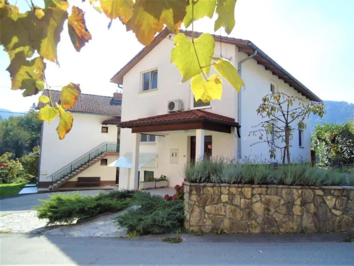 Bela hiša Gorjan z rdečo nadstrešnico nad vhodom, kamnitim zidom in vrtom, v ozadju hribi nad Tolminom